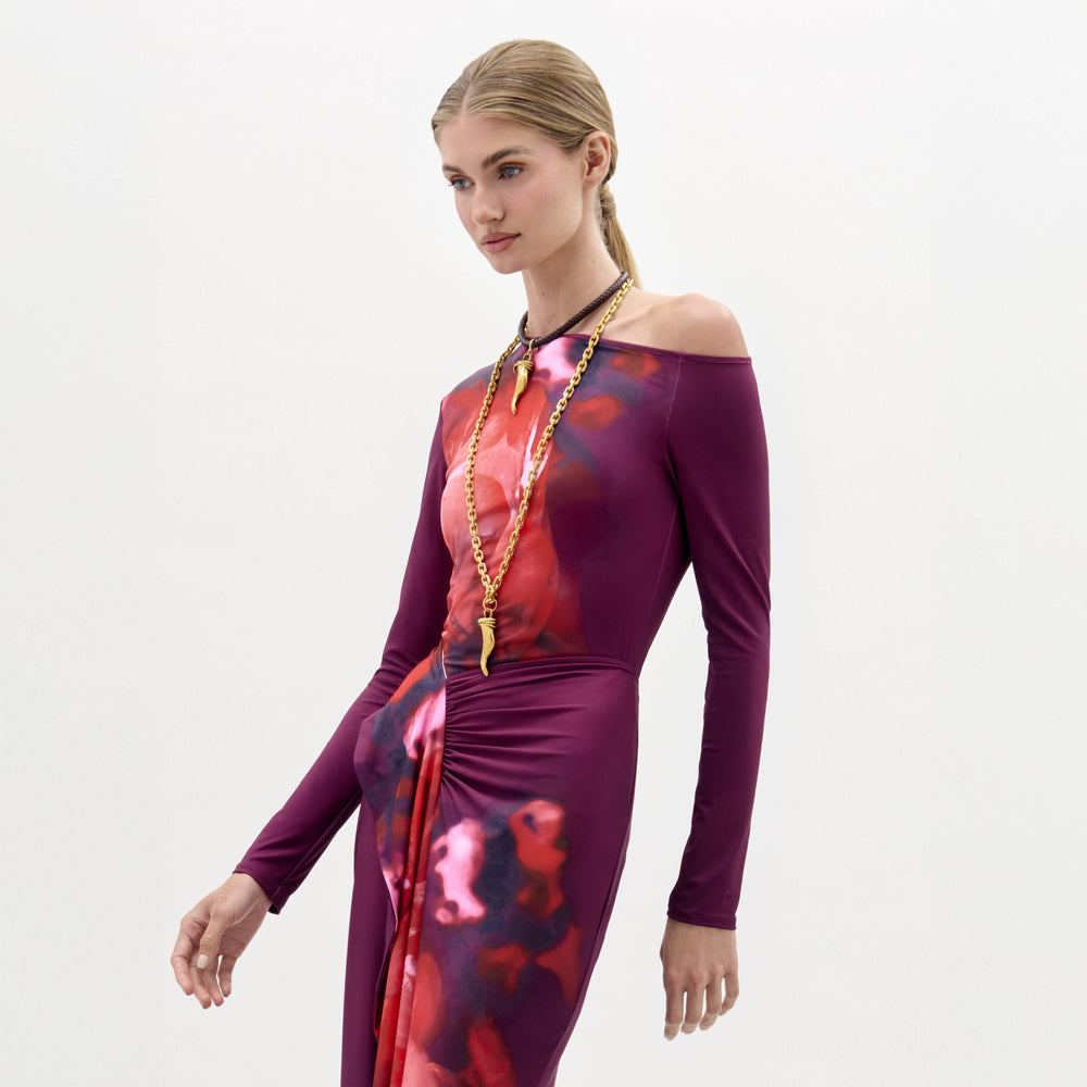 A woman wears the Sabra Blouse, a long-sleeve purple and red patterned top with an asymmetric neckline and a long gold necklace, standing against a plain white background.