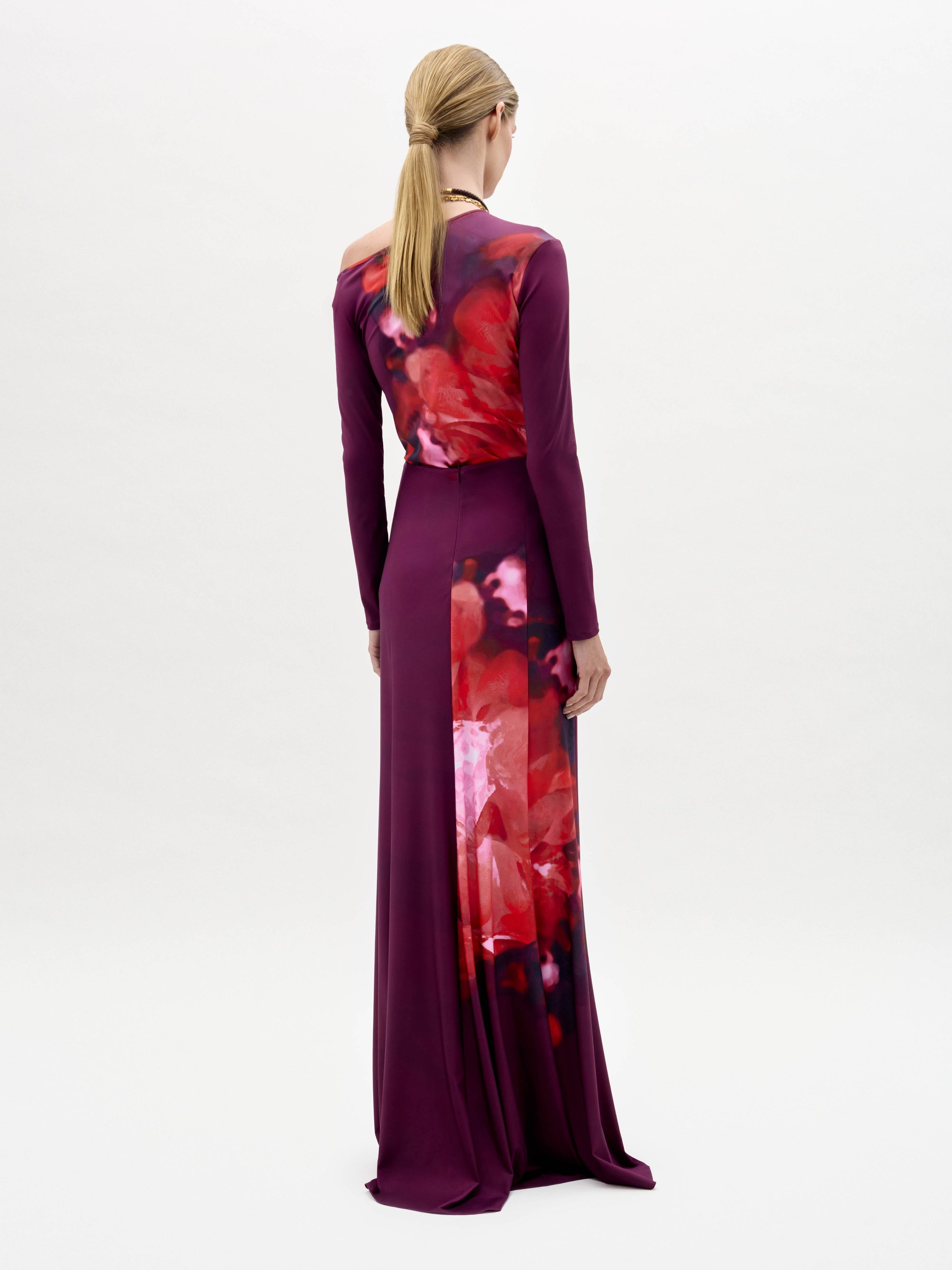 A woman models the Meissa Skirt, featuring a floor-length magenta design with a red and purple abstract pattern, paired with layered necklaces, against a plain white background.