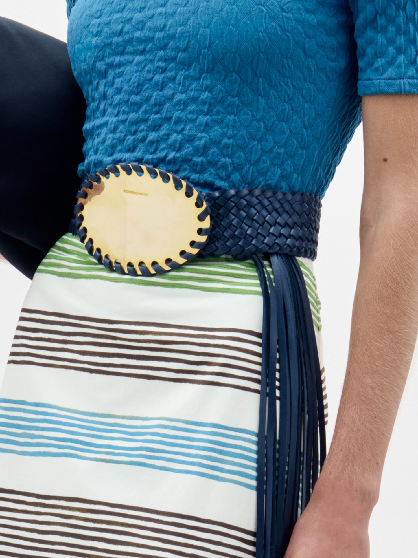 The Kai Belt is a black braided belt with a large oval gold buckle and long black fringe hanging down one side, shown on a plain light grey background.