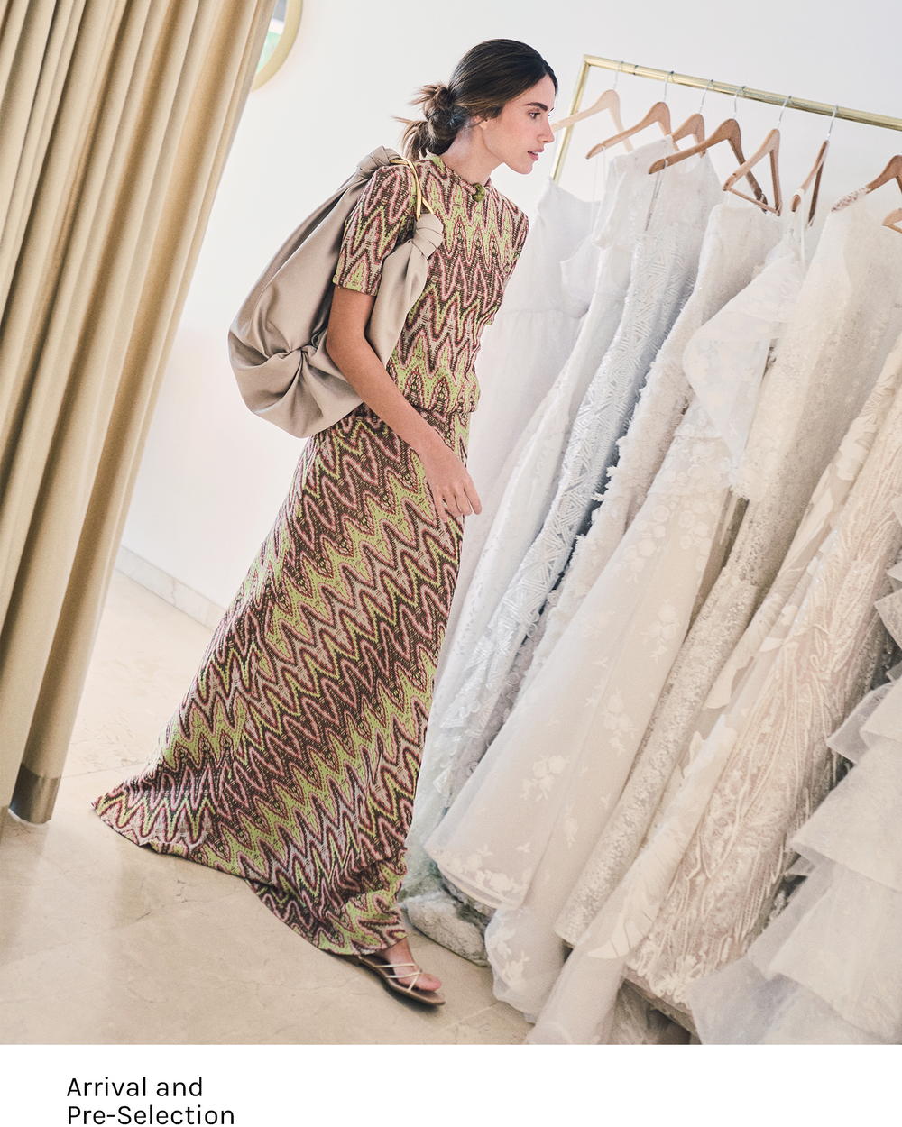 Woman in a patterned dress standing next to racks of white dresses in a fitting room.