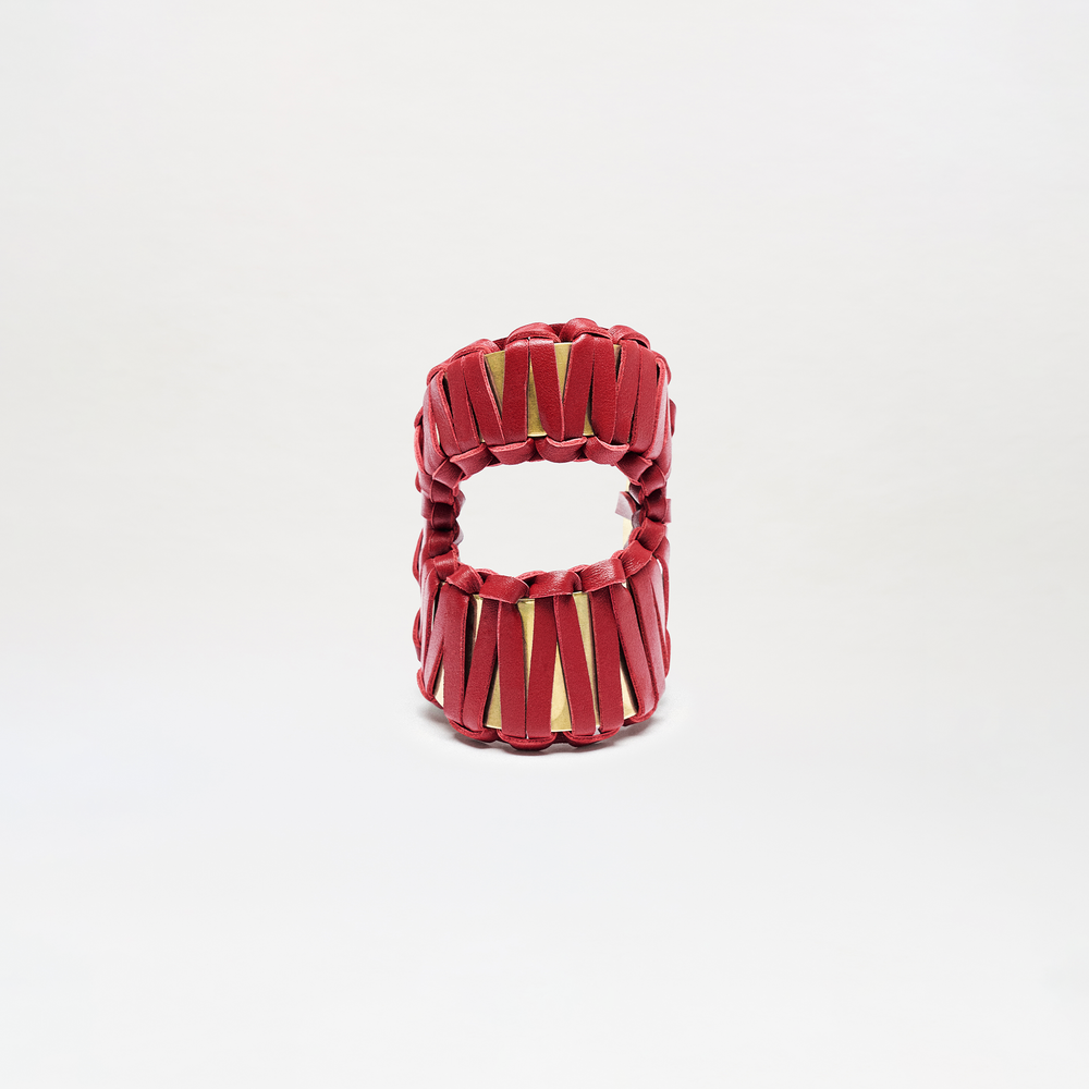 The Majel Cuff is a cylindrical accessory with a hollow center, featuring vertical red bands that crisscross and reveal a gold interior. It is displayed against a plain white background.