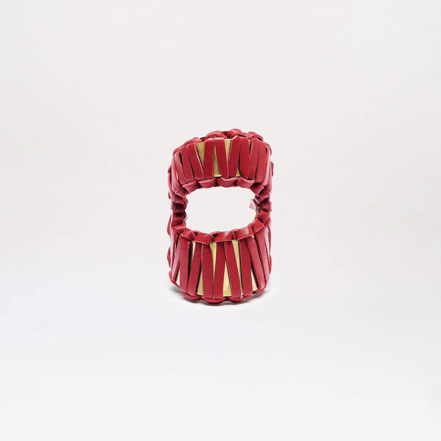 The Majel Cuff is a cylindrical accessory with a hollow center, featuring vertical red bands that crisscross and reveal a gold interior. It is displayed against a plain white background.