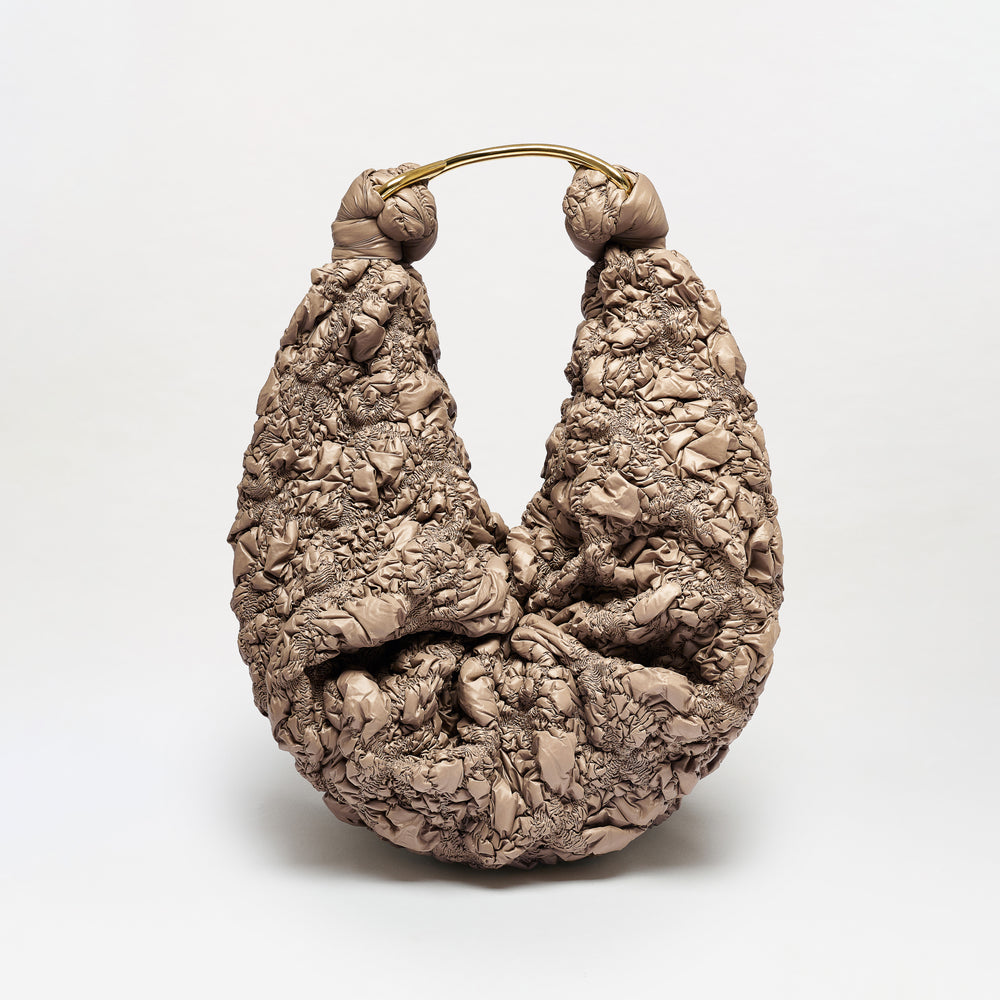 The Sofia Bag (Maxi) is a sculptural, crescent-shaped bag crafted from crinkled taupe Italian fabric with a curved gold-toned handle, shown standing upright against a plain white background.