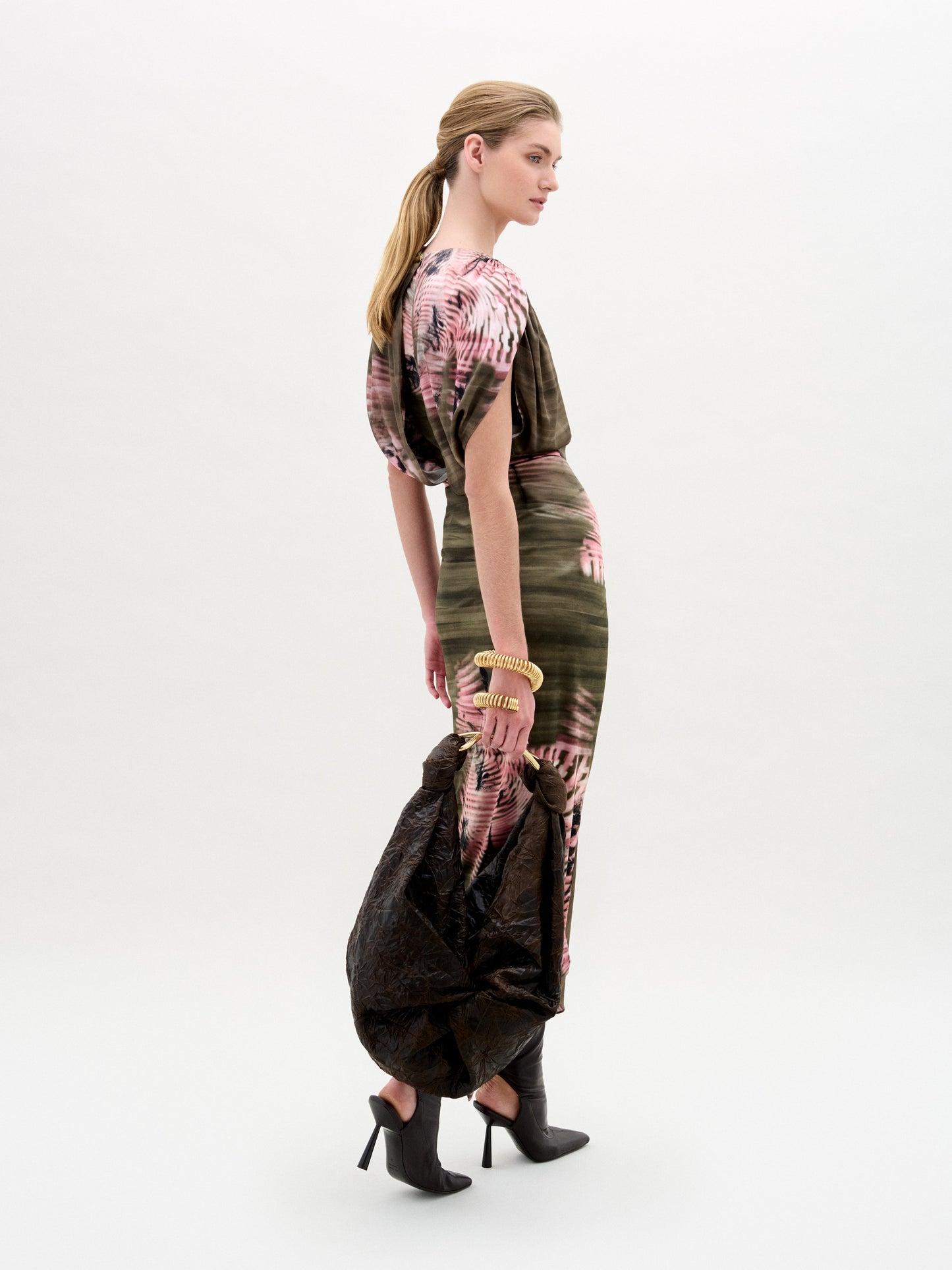 A woman stands against a plain background wearing a pink and brown patterned top, the Adrika Skirt in Italian fabric, black boots, and holding a large brown textured bag.