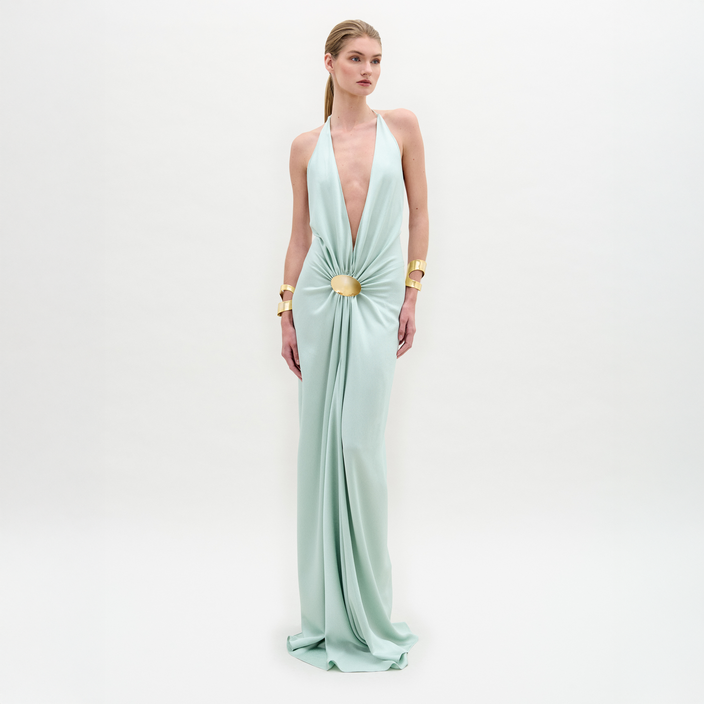 A woman wears the Aiko Dress, a light blue, floor-length maxi made from Italian fabric with a deep V-neck and central clasp, styled with gold bracelets against a plain white background.
