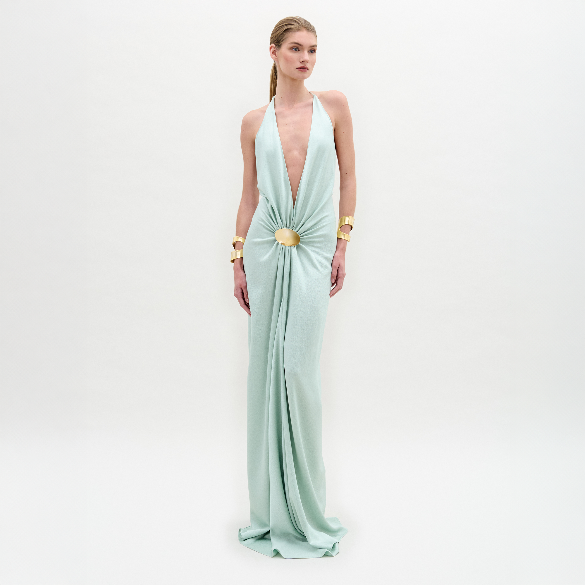 A woman wears the Aiko Dress, a light blue, floor-length maxi made from Italian fabric with a deep V-neck and central clasp, styled with gold bracelets against a plain white background.
