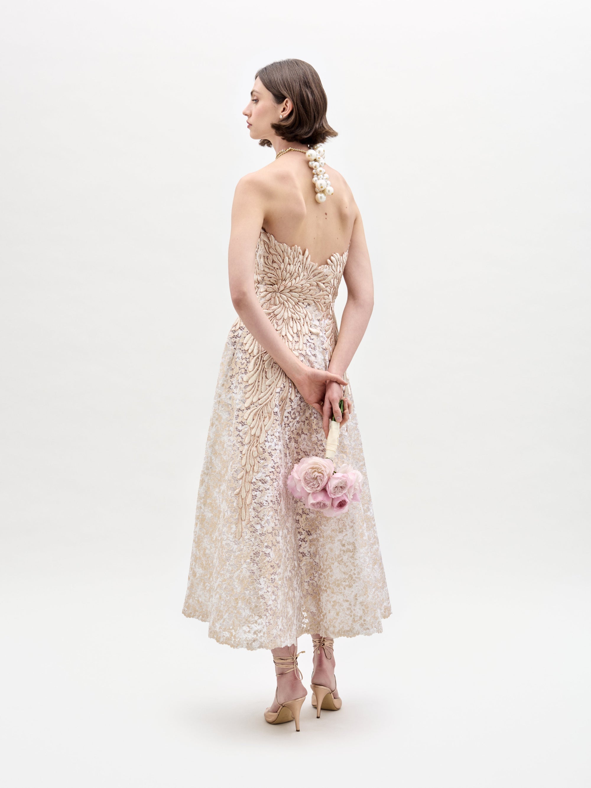 A woman in the strapless Alejandra Dress holds pink flowers. She wears pearl jewelry and beige heels, standing against a plain white background.