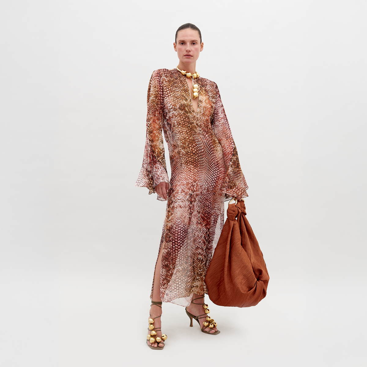 A woman stands against a plain background in the Aries Dress—a long, sheer abstract print made from 100% Italian silk in earthy tones. She carries a large rust bag, wears strappy gold-ball heels, and her hair is slicked back.