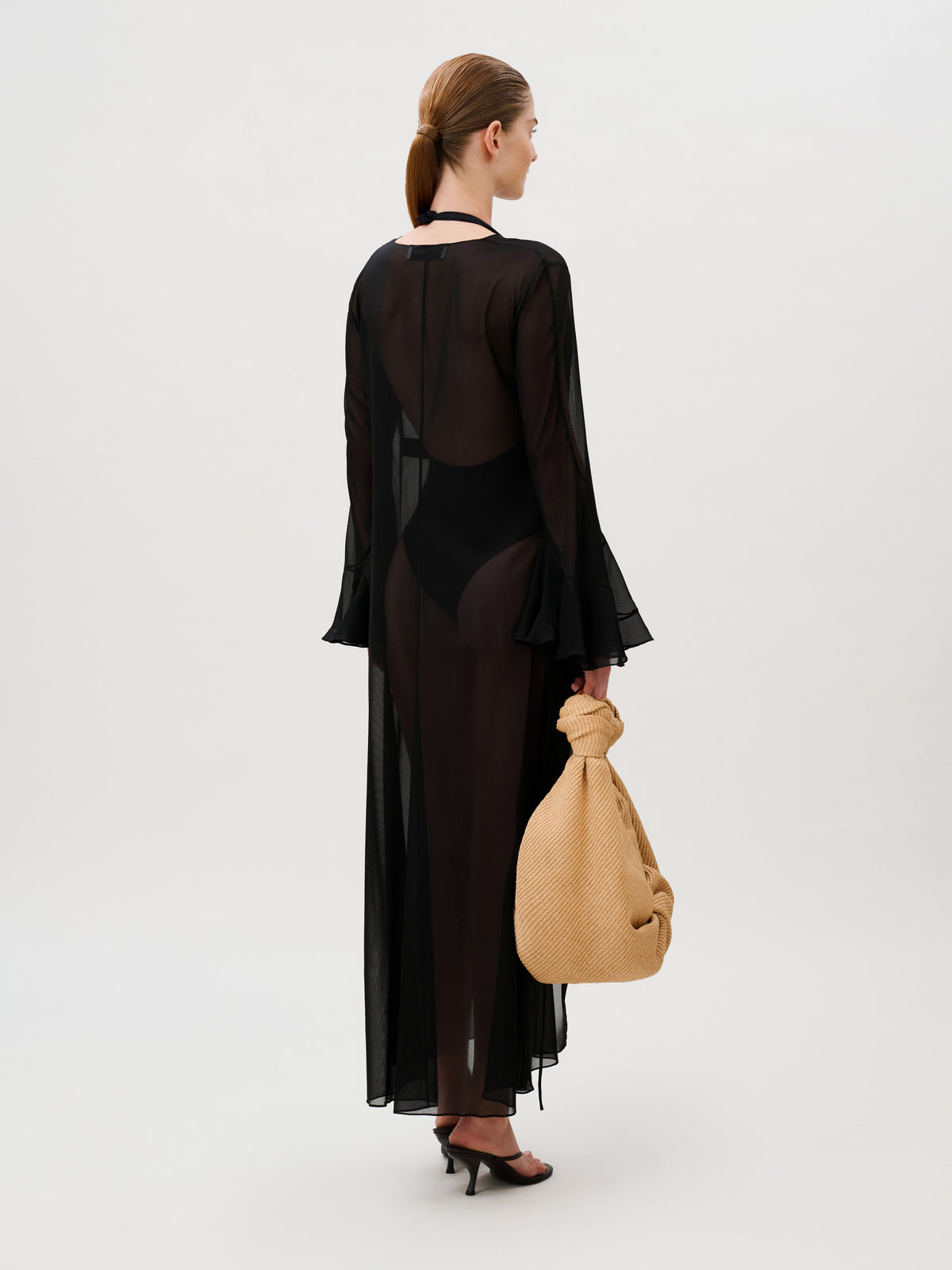 A person seen from behind wears a floor-length, sheer chiffon Amaris Tunic over a black swimsuit and holds a textured tan handbag.