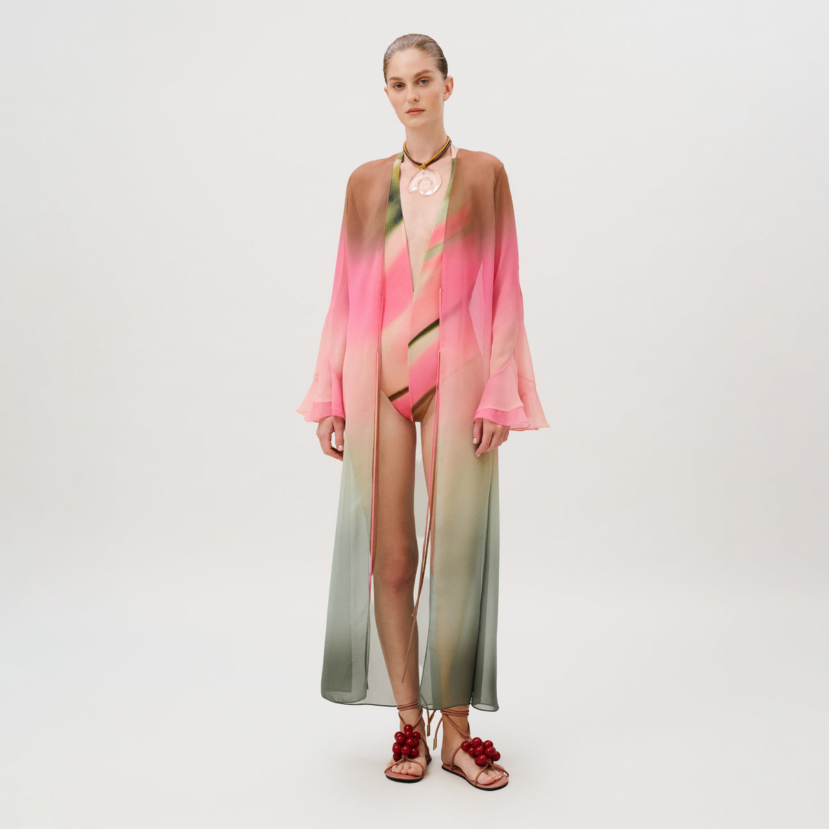 A model wears an Amaris Tunic, a floor-length, open-front chiffon kaftan with a pink, brown, and green gradient print over a matching bodysuit and tie-up sandals with red berry-like embellishments.
