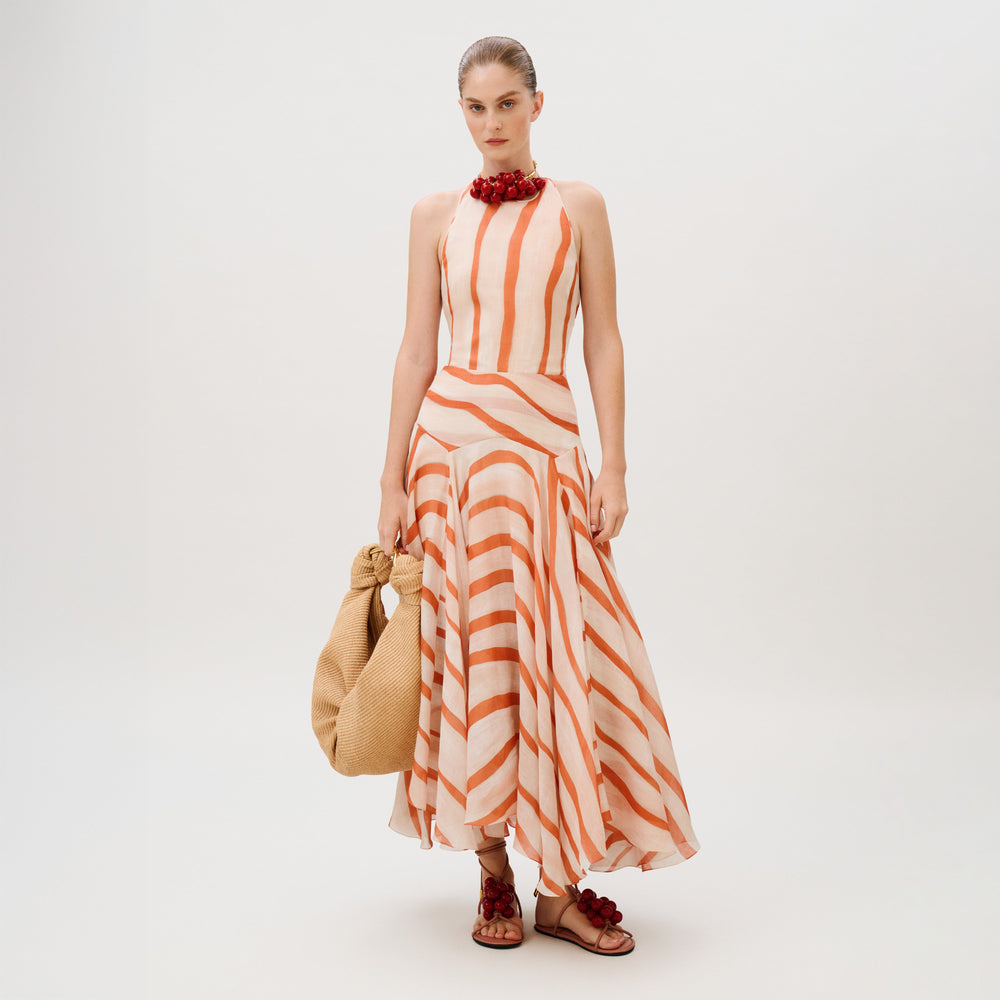 A model wears the Amity Dress, a sleeveless, halter-neck maxi in 100% cotton with orange and white diagonal stripes, paired with a woven tote bag and embellished flat sandals.