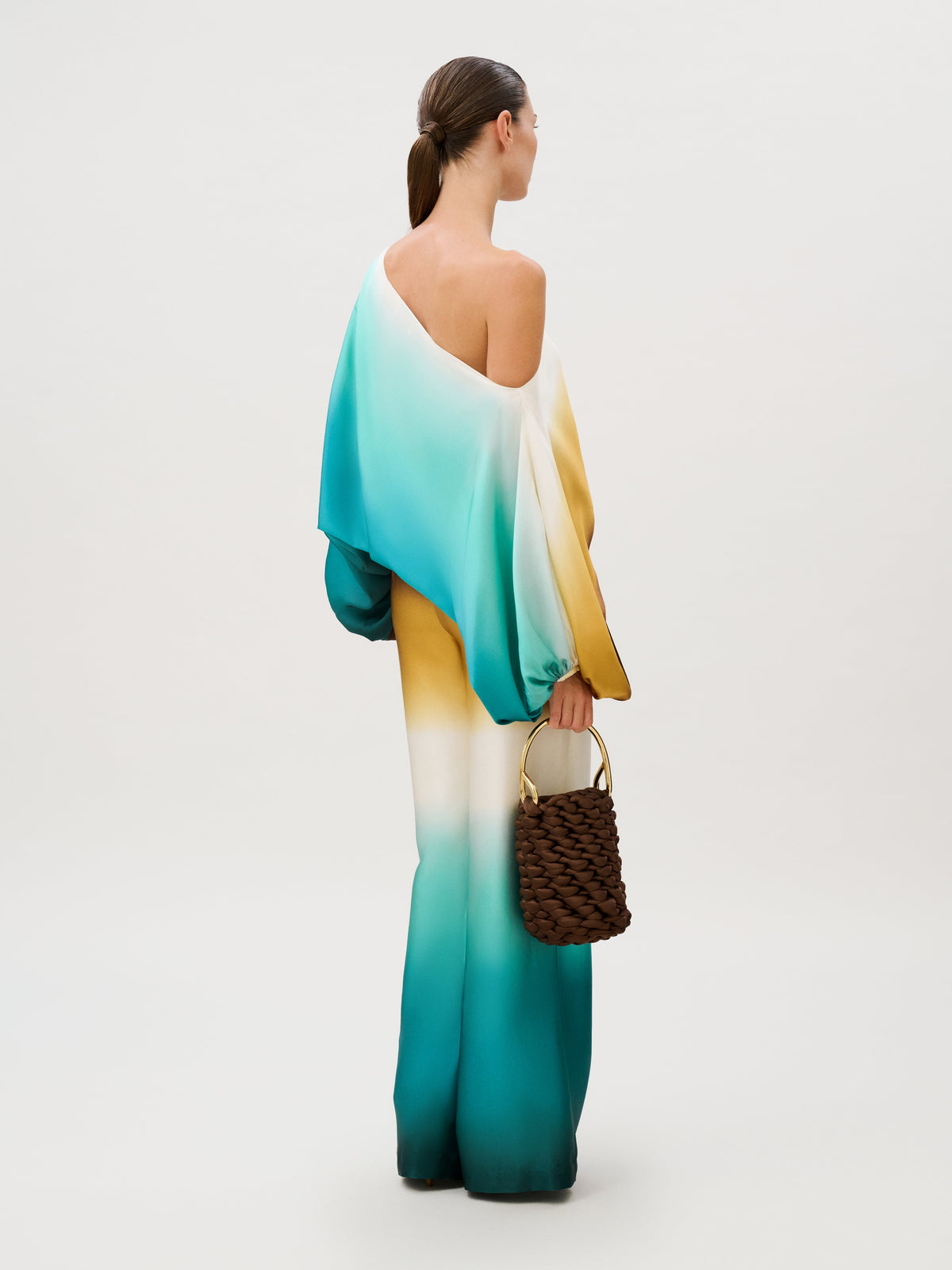 A person wearing an Andie Pant wide-leg one-shoulder jumpsuit with a white, yellow, and teal gradient, carrying a dark brown woven handbag with a gold handle.