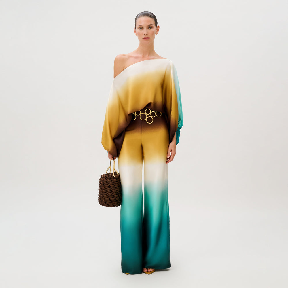 A person wearing an Andie Pant silk jumpsuit featuring an off-the-shoulder, ombre-patterned top in shades of gold, brown, and teal, cinched with a gold-ringed belt and holding a dark brown woven handbag.