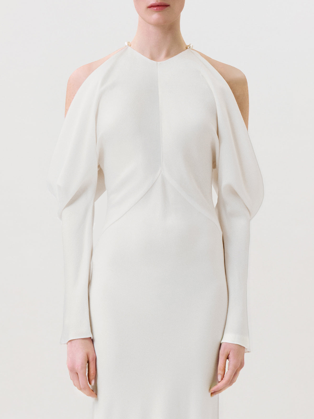 A person wearing the Antonia Dress, a sleek, long-sleeved, white hammered satin garment with open shoulders and a high neckline. The ready-to-wear dress is fitted and elegant, shown against a plain white background.