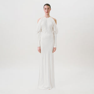 A person stands facing forward wearing the Antonia Dress—an elegant, long white gown featuring cut-out shoulders, long sleeves, and a fitted silhouette crafted from luxurious hammered satin—all set against a plain white background. This Ready To Wear piece exudes modern sophistication.