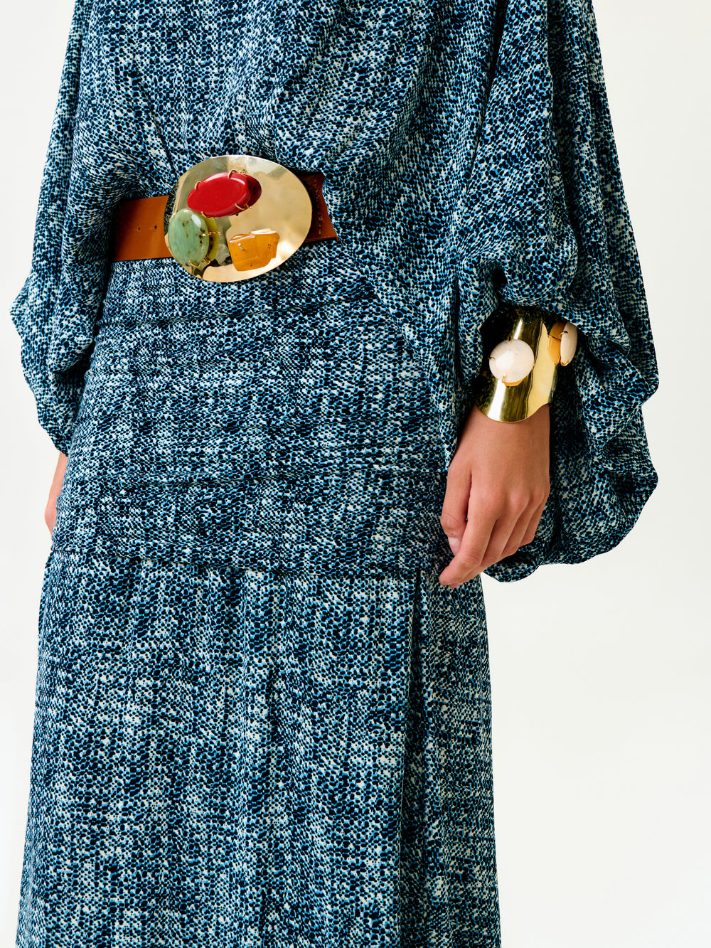 A person wears the Nelly Dress—a textured blue and black patterned maxi with loose sleeves—paired with a wide brown belt featuring a bold, multicolor buckle and a chunky gold bracelet. Made in Colombia; shown from shoulders to knees.