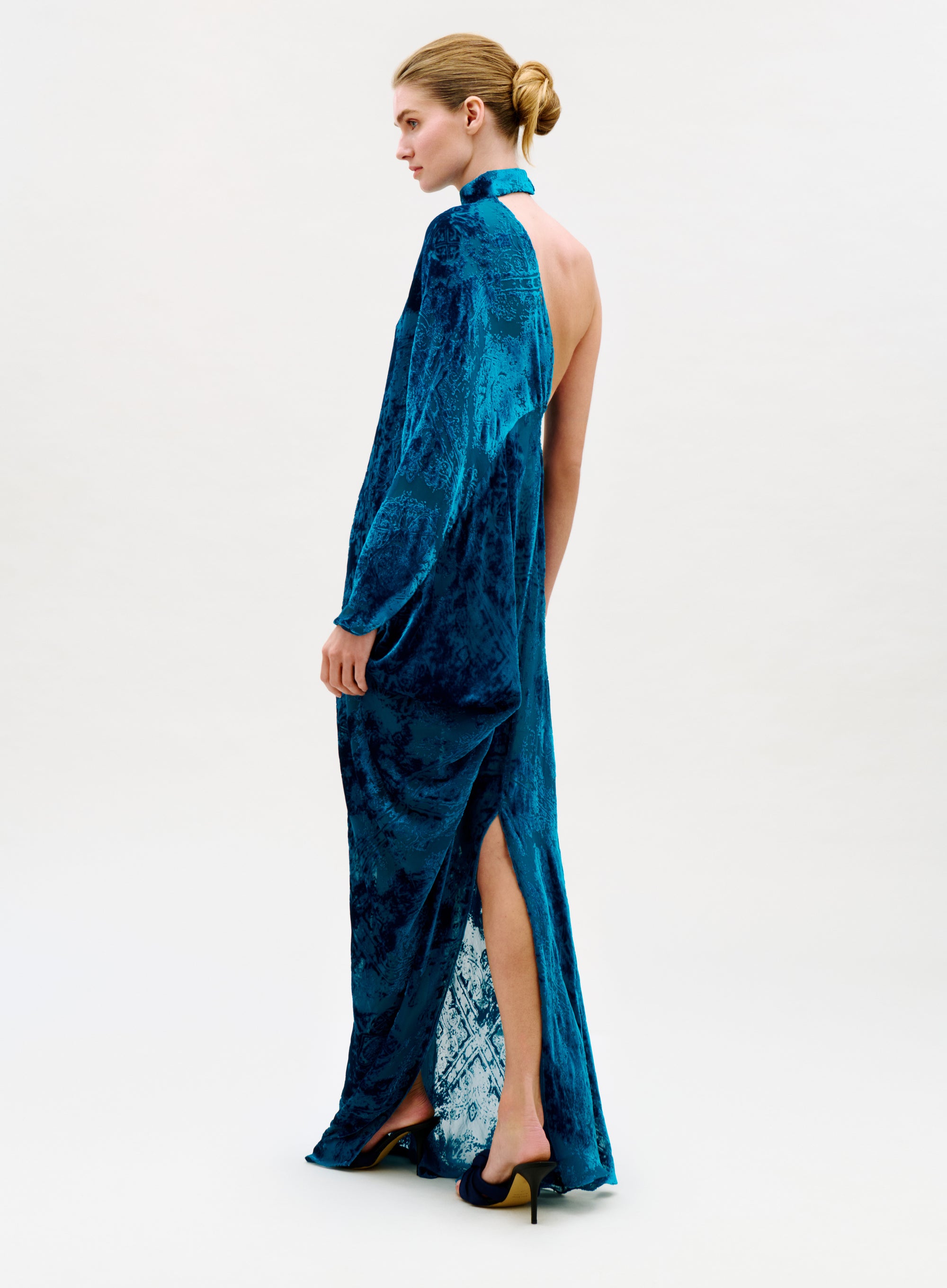 A woman in a low bun showcases the Anya Dress, a long blue velvet gown with an open back and high slit, paired with black heels against a plain light background.