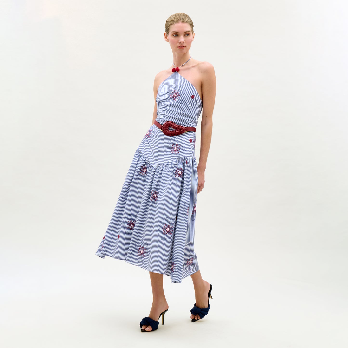 A woman stands against a plain white background wearing the Ares Dress, a light blue halter-neck midi dress with red floral patterns, an A-line silhouette, a red belt, and dark heeled sandals.