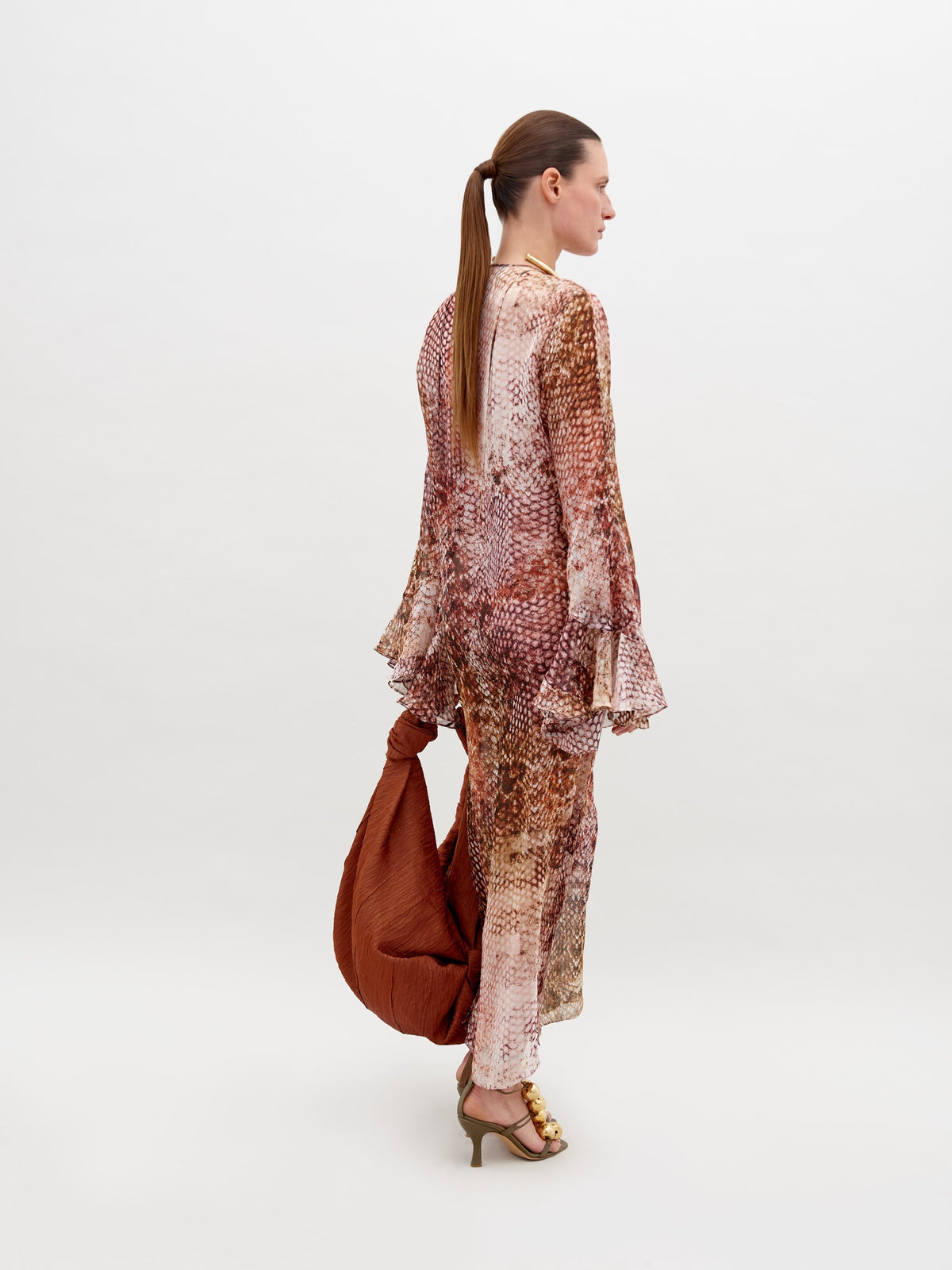 A woman with a low ponytail wears the Aries Dress—a long, 100% silk abstract print dress with flared sleeves—paired with gold heeled sandals and a large brown textured bag against a plain white background.