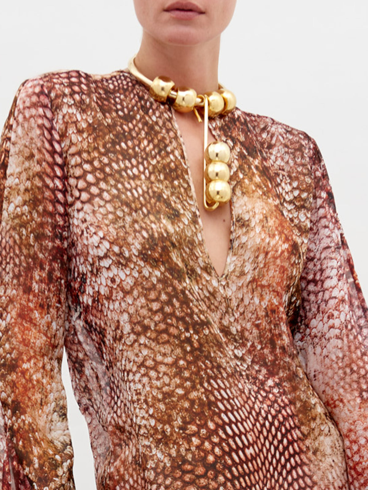 A person wearing the Aries Dress, a v-neck, long-sleeved 100% silk dress with a brown and cream snakeskin pattern, paired with a chunky gold bead necklace. Only their lower face and upper torso are visible.
