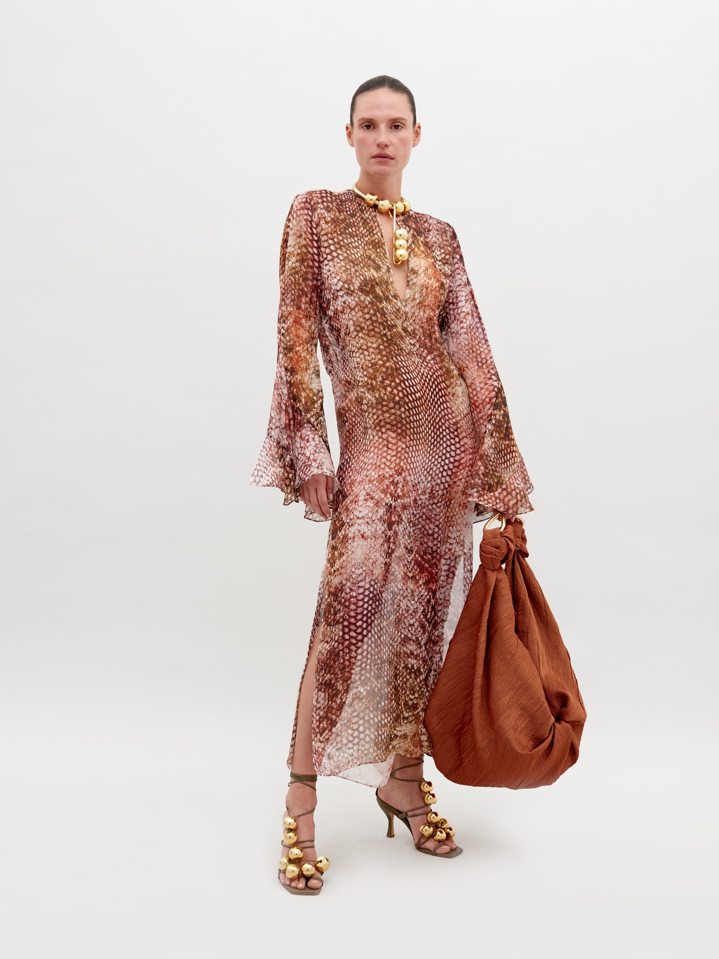 A person stands against a plain background wearing the Aries Dress Peach Animal Print with flared sleeves, embellished accessories, and high-heeled sandals. They hold a large, rust-colored bag. Pre-order now to have it by November 15th, 2024.