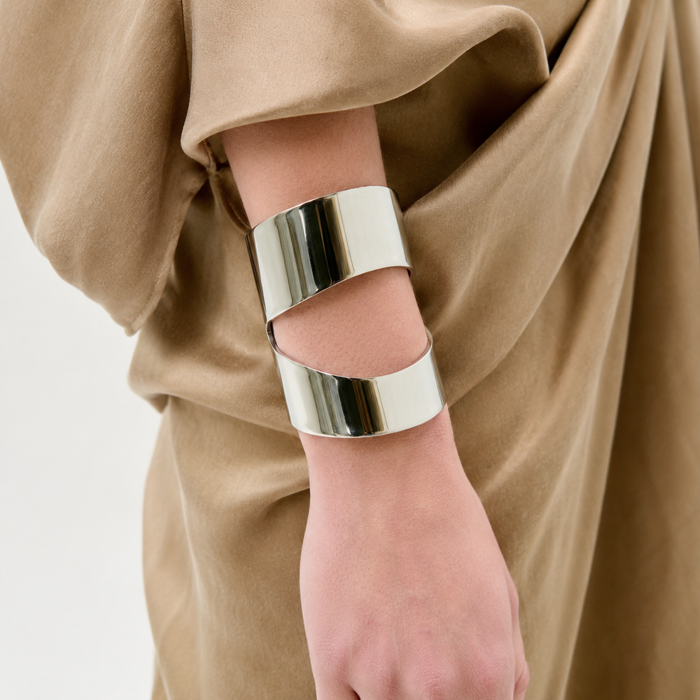 A person wears a camel-colored draped garment and the Artemis Cuff—a wide, modern silver bracelet with a unique overlapping open design—on their wrist.