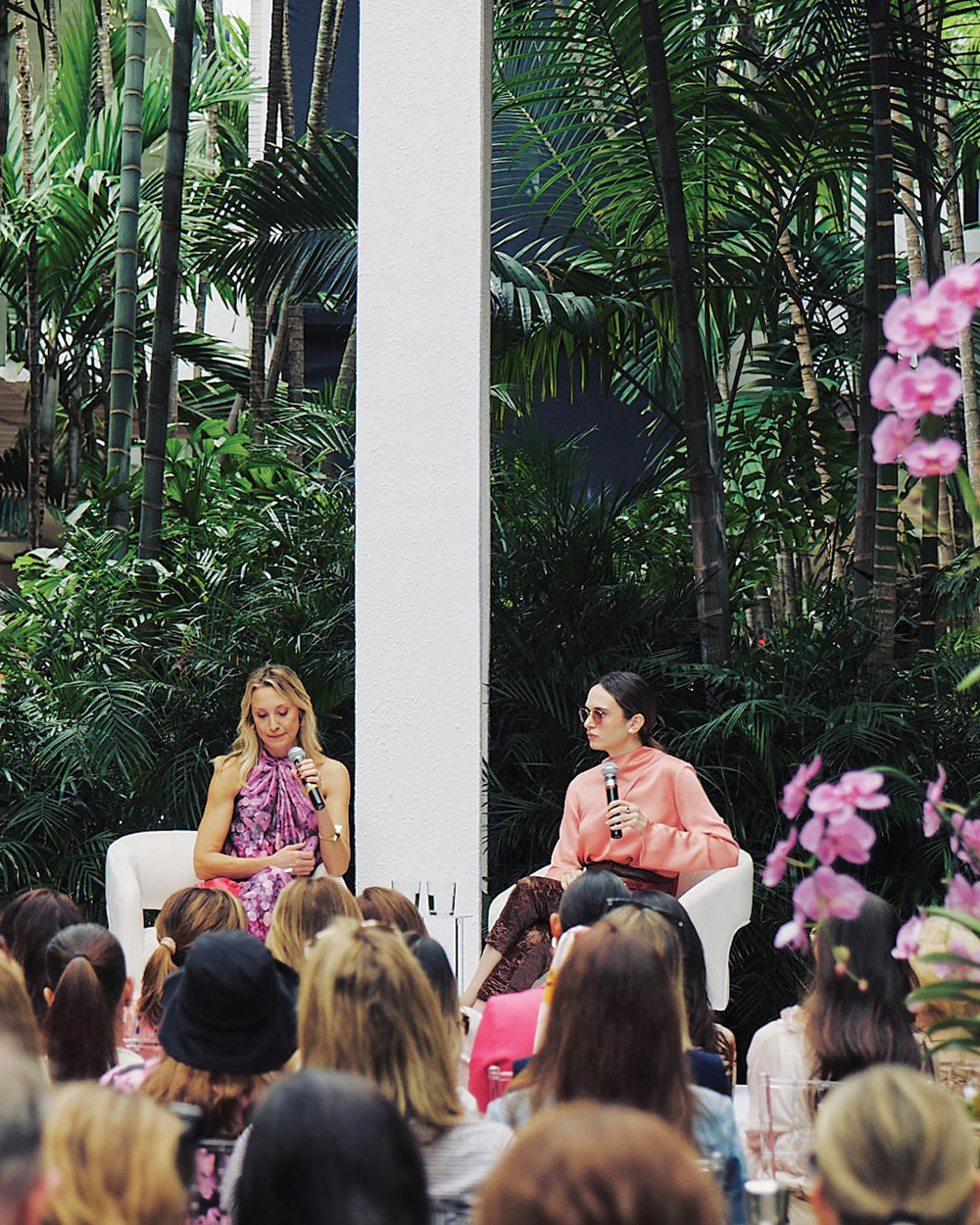 Two women speaking at a podium with a tropical setting and audience in the foreground.