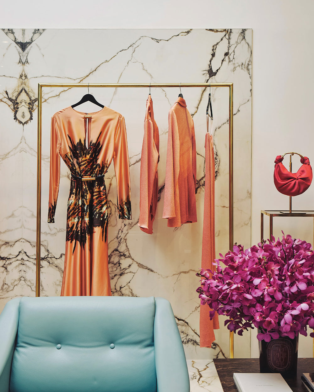 Orange dress with black detailing hanging on a marble wall with a blue chair and pink flowers in the foreground.