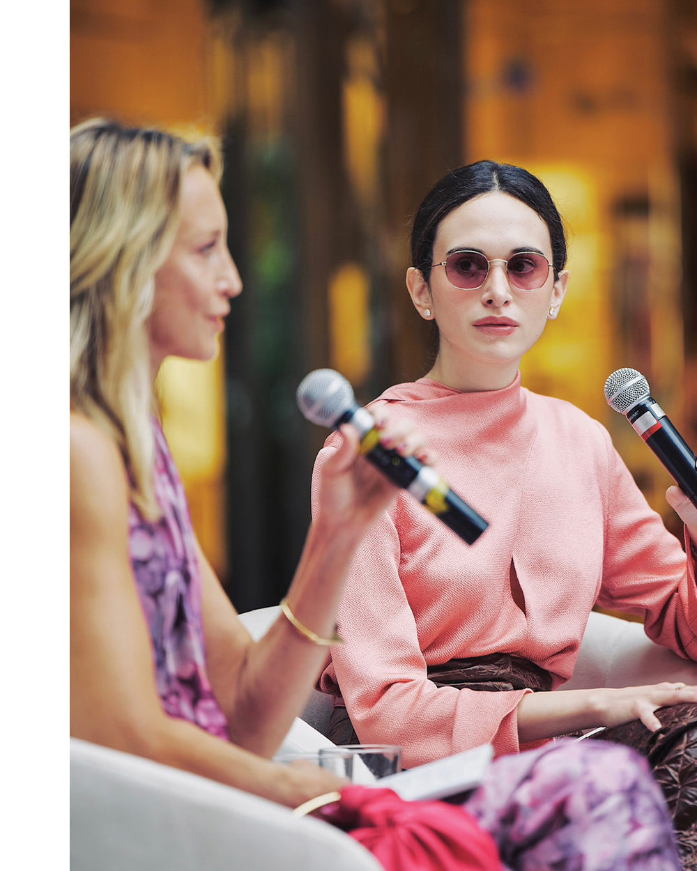Woman in pink sweater and sunglasses holding a microphone, with another person in a purple top beside her.