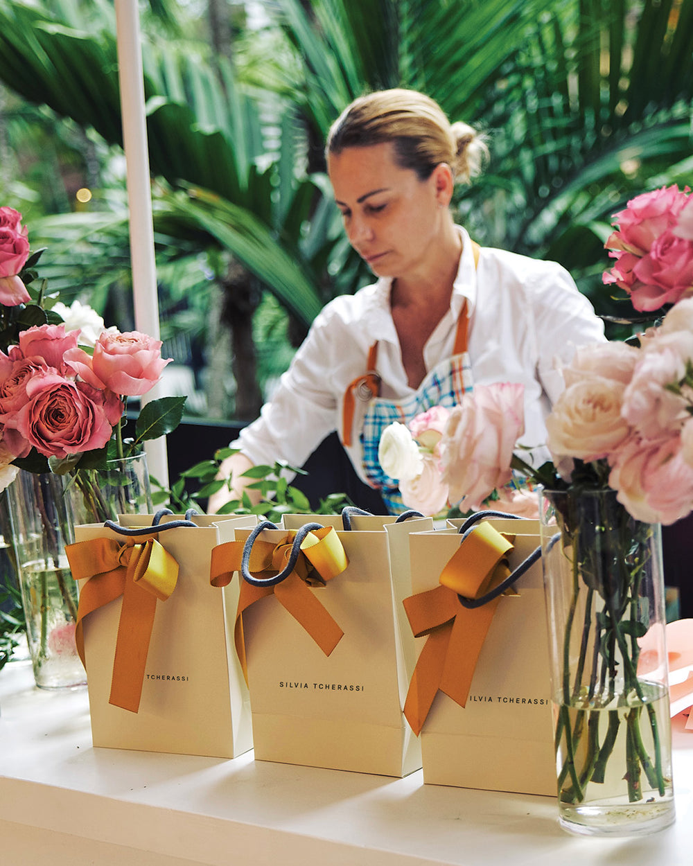 Woman arranging flowers with gift bags and vases on a table outdoors.