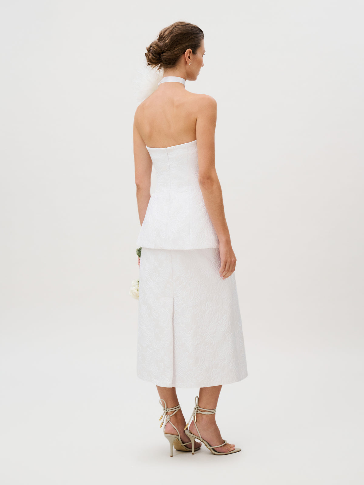 The woman wears the Atira Skirt, designed in a luxurious Italian white fabric with a strapless, halter neckline. She stands with her back to the camera, pairing the skirt with strappy heels and an elegant updo against a plain, light background.