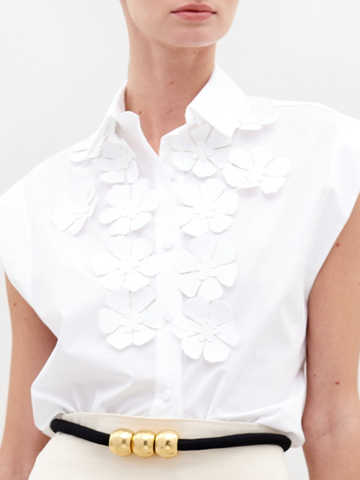 A person wearing the Azalea Blouse in white, which is sleeveless and adorned with appliqué floral detailing, paired with a skirt featuring a black belt and gold accents.