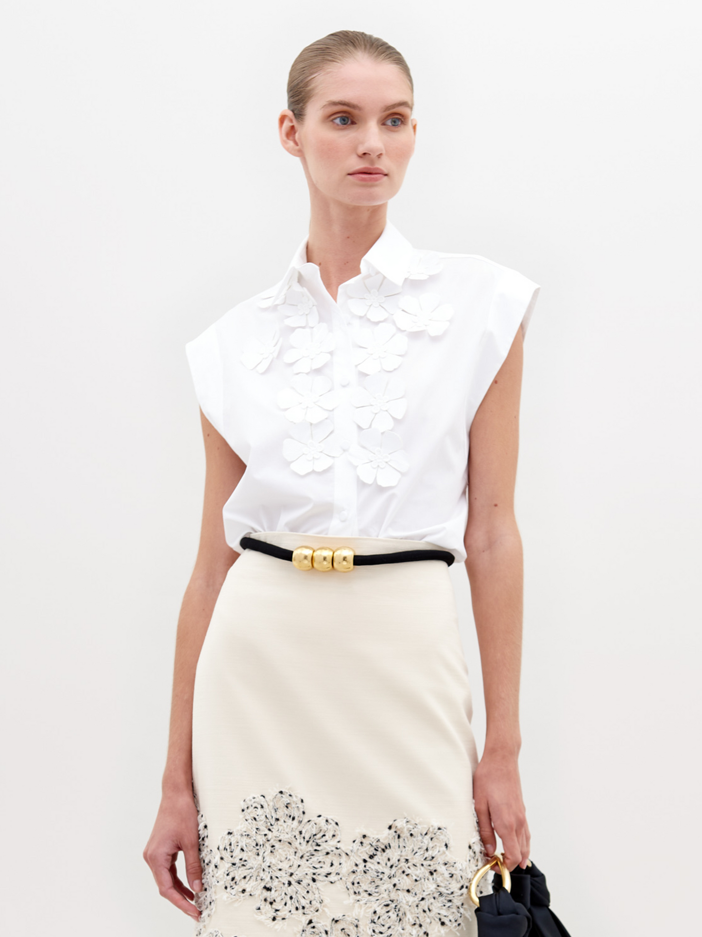 A person wearing the Azalea Blouse, a sleeveless white top crafted from Italian textile with floral detailing, and a beige skirt adorned with embellishments, stands against a plain background.