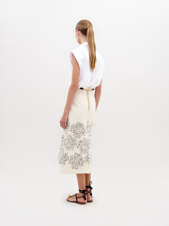 A woman stands wearing a white patterned jacket, Atira Skirt Beige Black Embroidered Flowers, gold belt, and gold-embellished sandals. She holds a beige handbag against a plain background. Don't miss out—pre-order now for the November 15th, 2024 ship-by date!