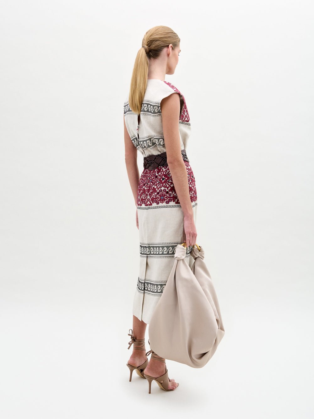 A woman stands against a white background wearing the Badra Dress, styled with a woven leather belt, beige strappy sandals, and holding a large beige bag.