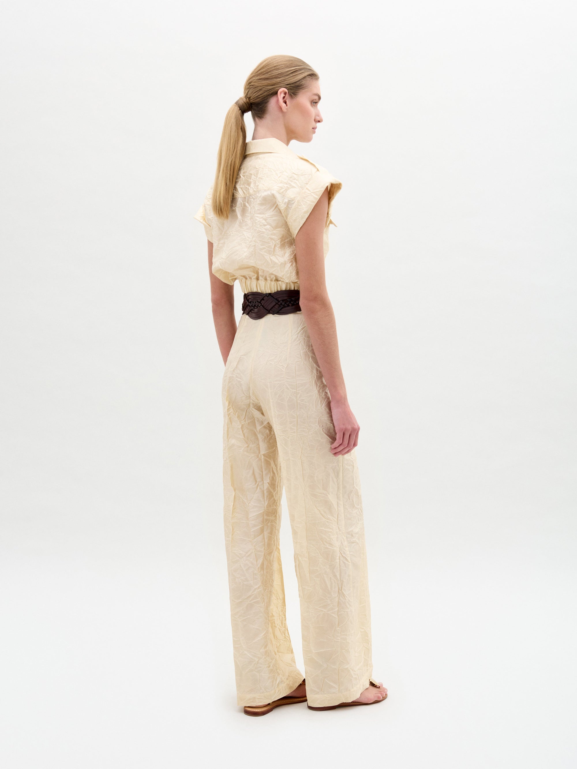 A person stands against a plain background wearing the Emine Pant, a beige textured wide-leg silhouette crafted from Italian fabric, styled with short sleeves, a wide black belt with gold buckle, and sandals.