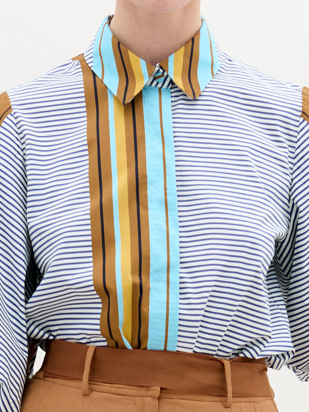 A person stands against a plain background wearing the oversized fit Belkis Blouse with balloon sleeves and stripes, paired with tan high-waisted pants, and holding a tan accessory in their left hand.