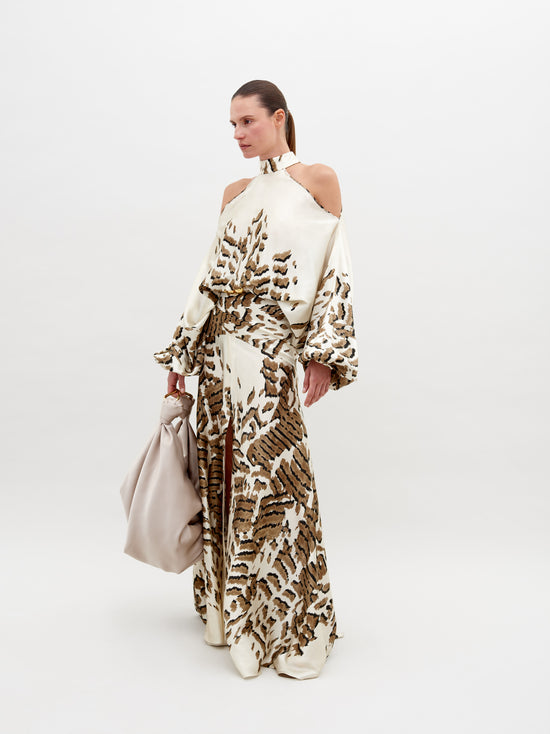 A woman poses in a beige and brown patterned, off-shoulder Bahar Dress Beige Abstract Animal Print holding a large, light beige handbag against a plain white background. This stunning ensemble, available for pre-order now, promises an elegant addition to your wardrobe with an estimated ship date in 2024.