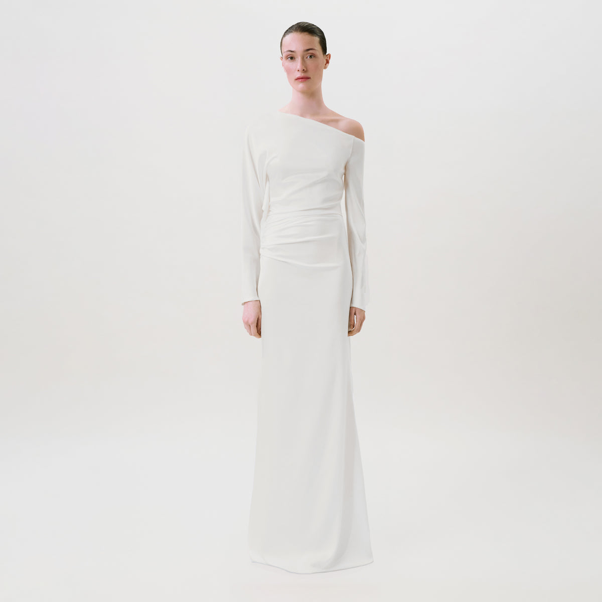A woman stands against a plain white background wearing the Bahir Dress, a long, white, one-shoulder gown crafted from luxurious silk. This elegant ready-to-wear piece features long sleeves and a draped waist detail, expertly made in the USA for effortless sophistication.