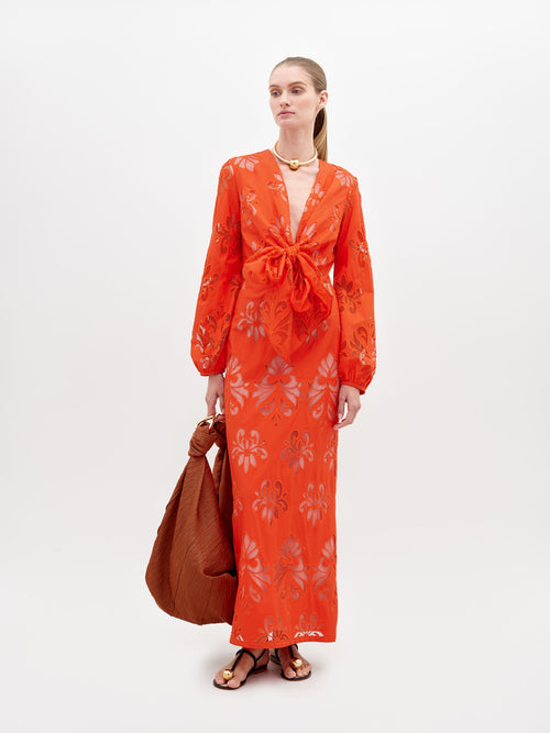 A person stands in a long, bright orange Battia Dress Rouge Embroidery with floral patterns and a front bow tie. They carry a large, rust-colored bag and wear gold jewelry and black sandals. To ensure you receive this stunning ensemble by the desired ship-by date, consider placing your pre-order for 2024 today.