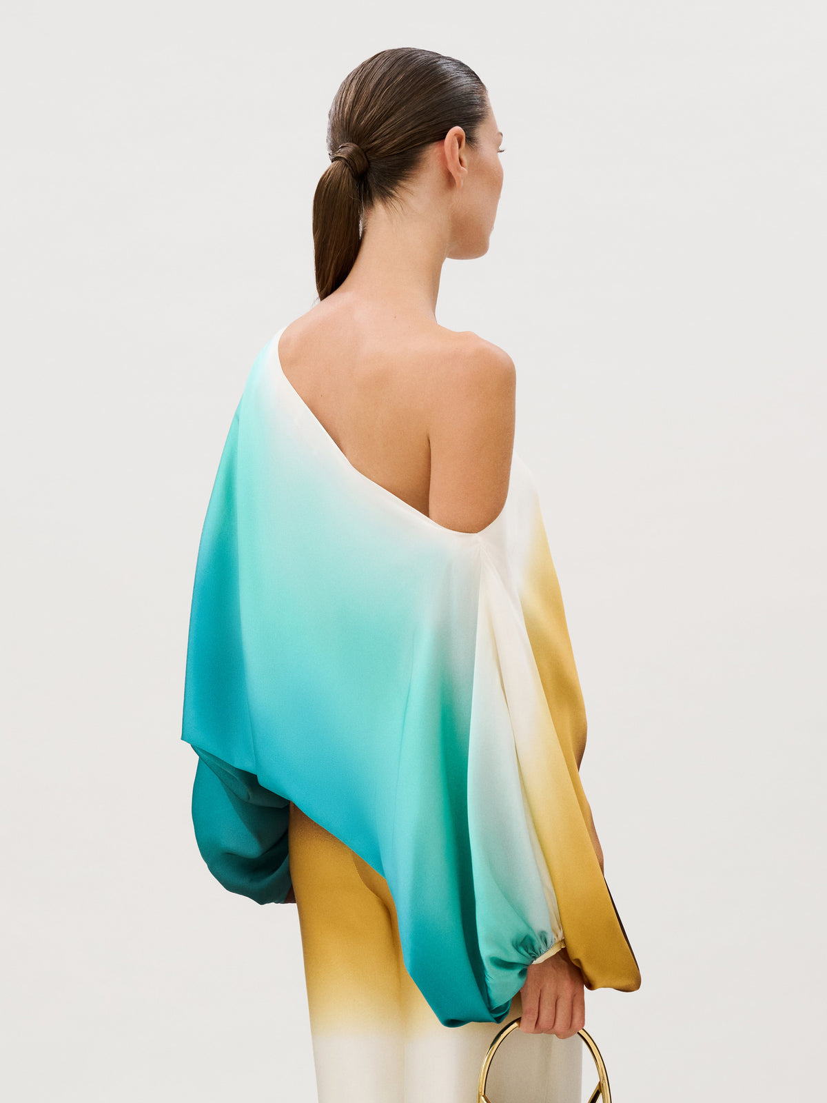 The Bellagio Blouse is an Italian silk one-shoulder top with a flowing, ombre design that transitions from teal to white to yellow, worn with matching wide-leg trousers.