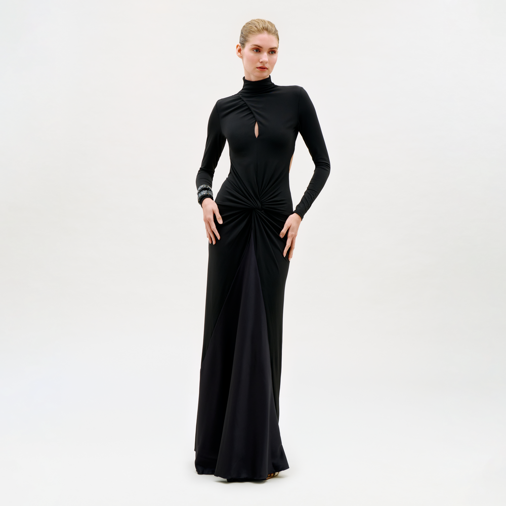 A woman wears the Birdy Dress—a long-sleeved, floor-length black gown featuring a small front keyhole cutout and gathered waist—standing against a plain, light background.