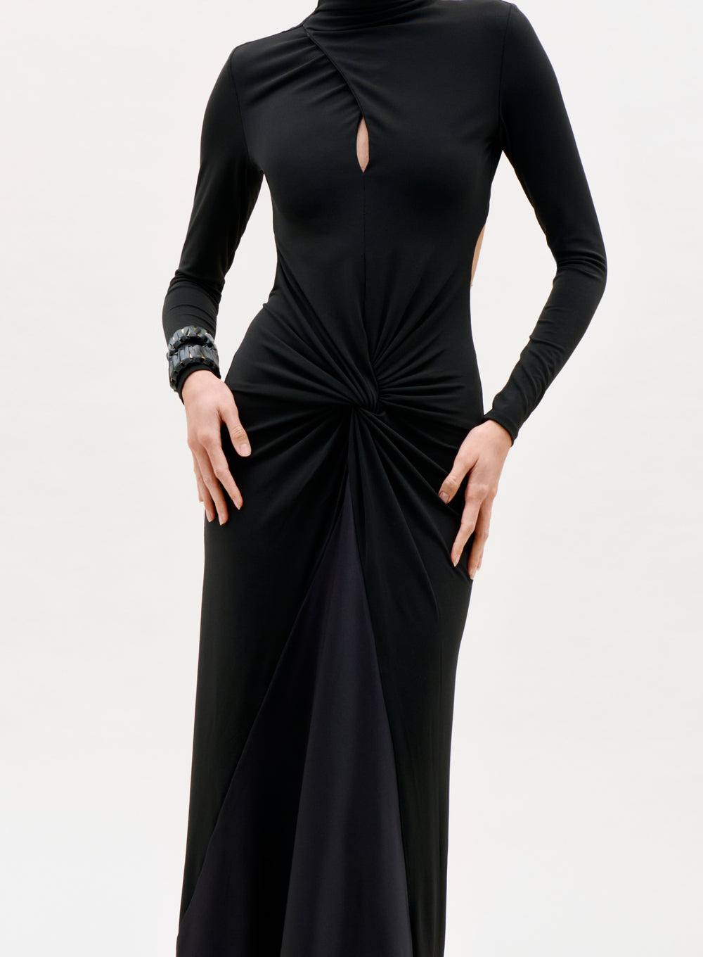 A person wearing the Birdy Dress, a long-sleeved black dress featuring a keyhole chest cutout and gathered twist at the waist, with a bracelet on one wrist, stands against a plain light background.