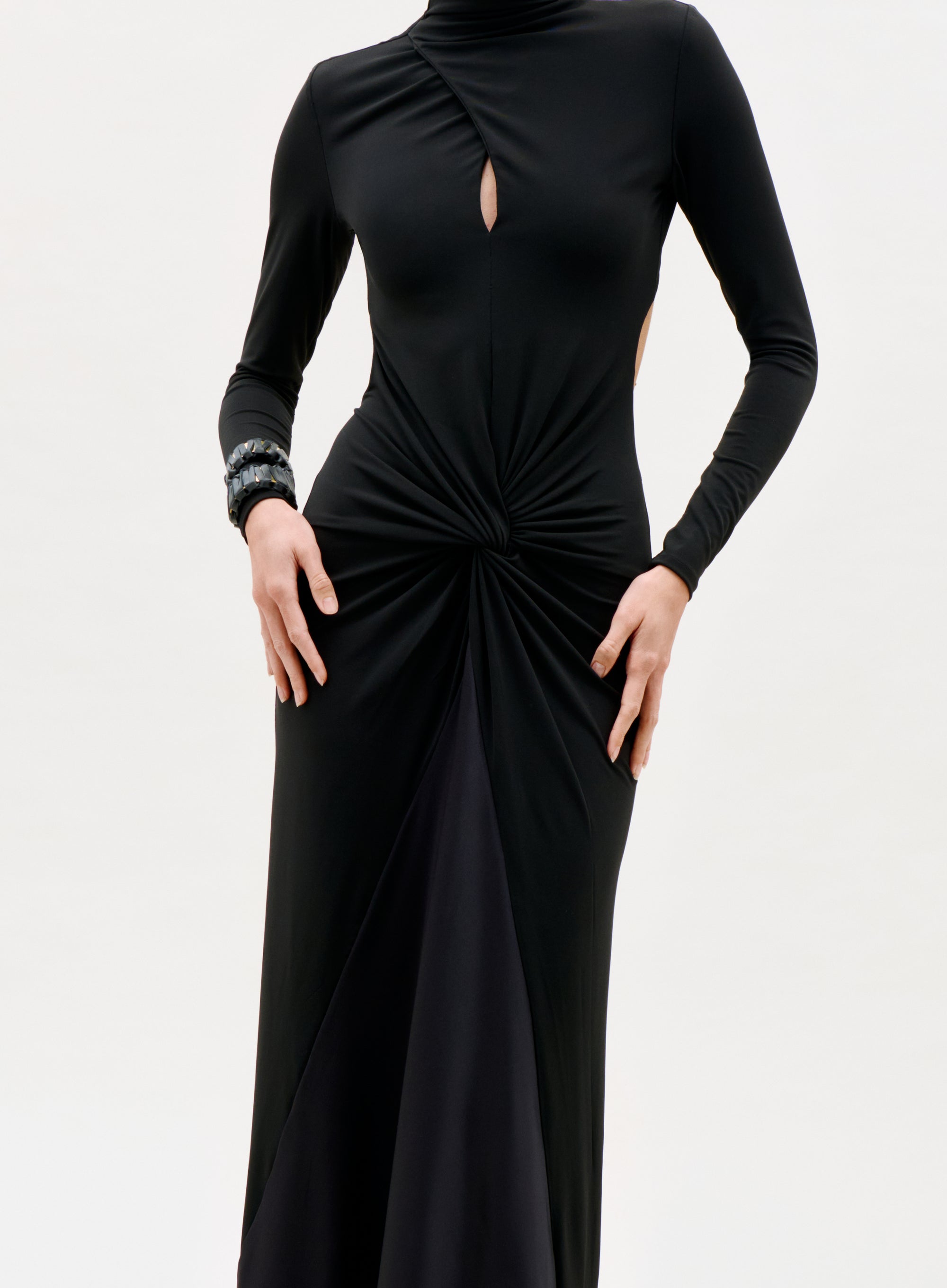 A person wearing the Birdy Dress, a long-sleeved black dress featuring a keyhole chest cutout and gathered twist at the waist, with a bracelet on one wrist, stands against a plain light background.