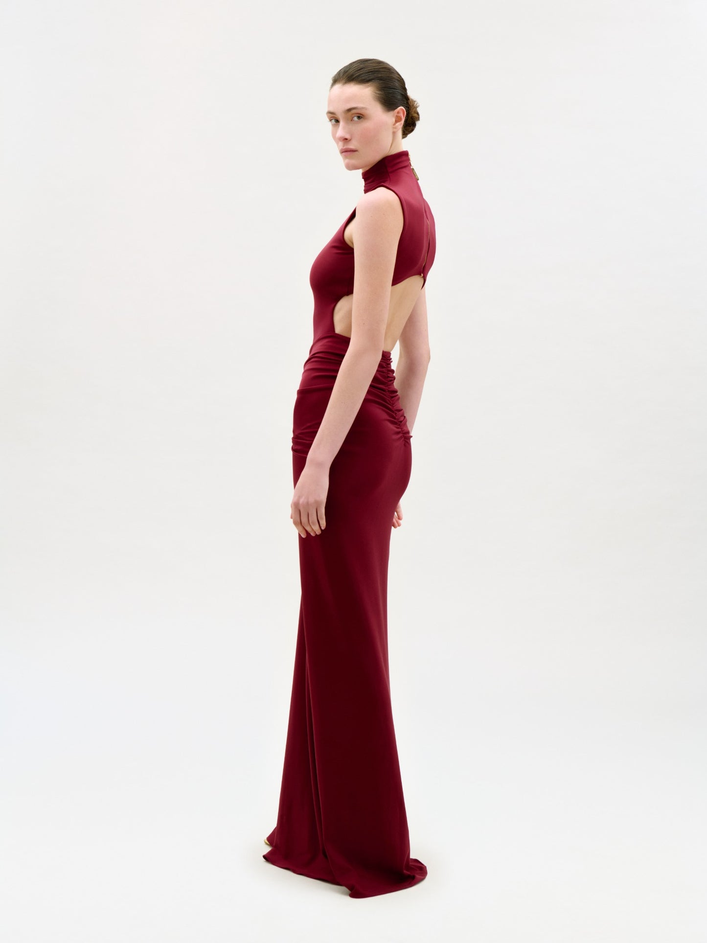 A woman stands sideways in the Bogna Dress, a sleeveless, high-neck, backless gown in dark red that drapes to the floor. The plain white background highlights the elegant, fitted silhouette.