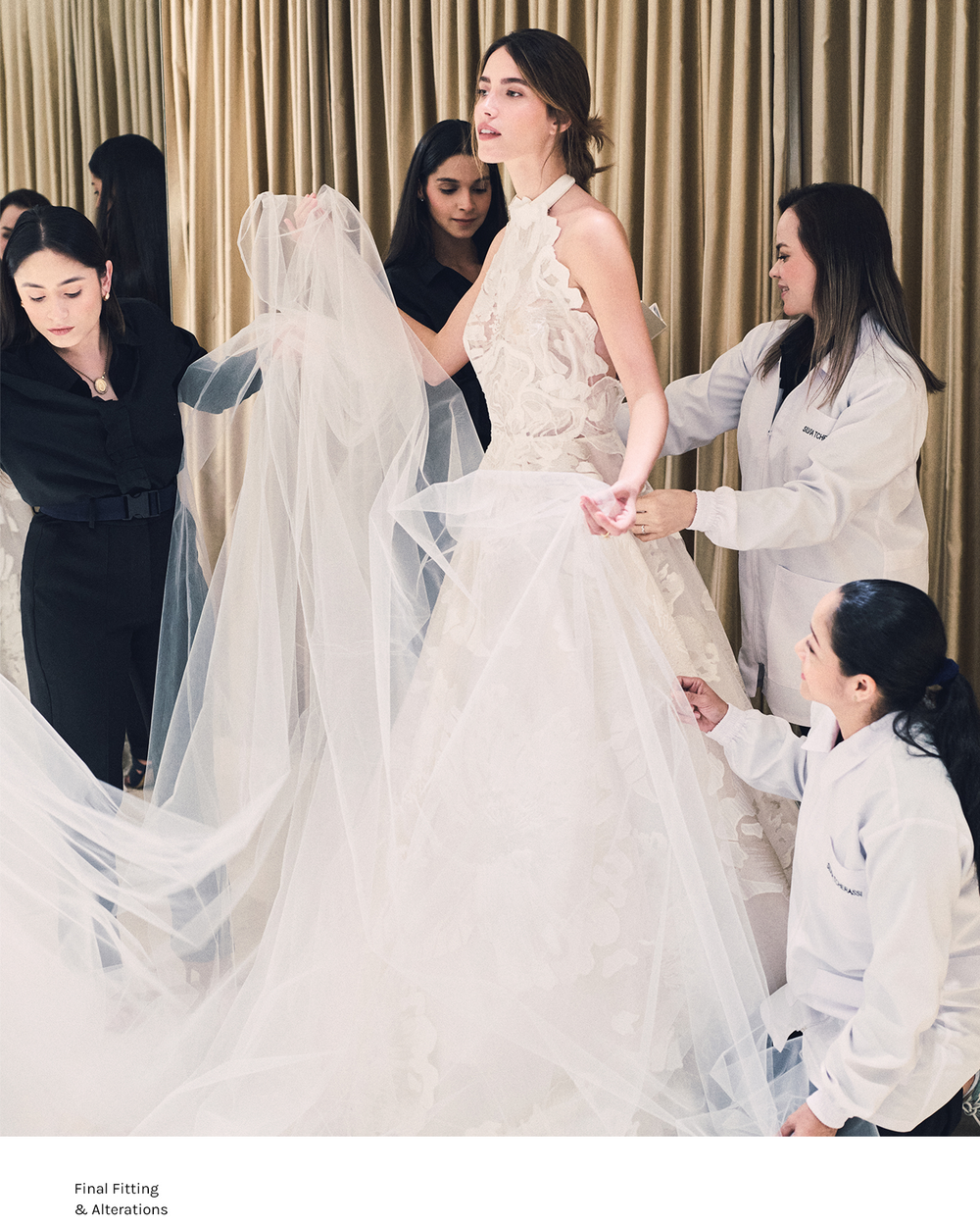 Model in a white wedding dress with a veil being fitted by professionals against a beige curtain background.