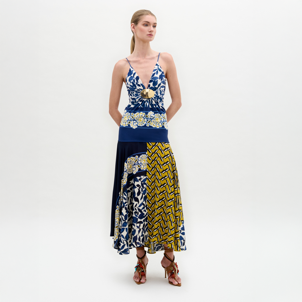A woman poses against a white background wearing the Casiana Dress, a sleeveless blue, yellow, and white patterned midi with a flared jersey skirt, styled with brown strappy sandals.