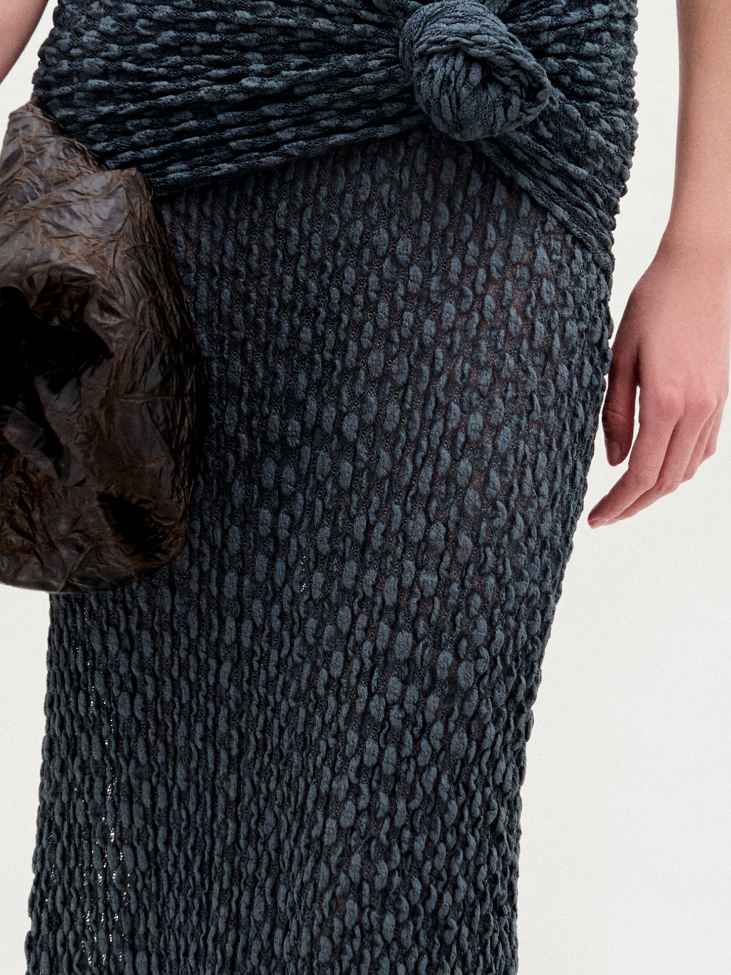 Wearing the Ciji Skirt—a form-fitting, textured dark knit midi with a knotted waist detail—a person holds a crinkled dark brown bag, showing only their lower torso and one hand against a plain background.