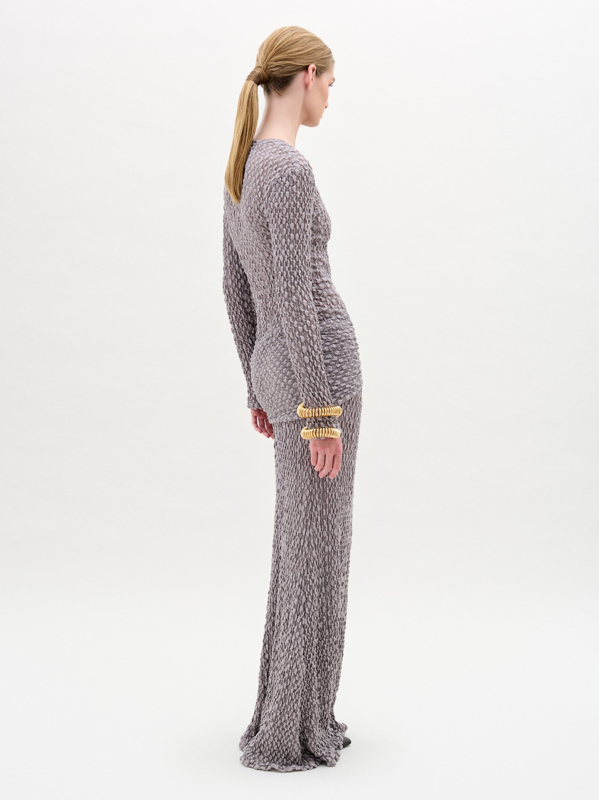A person with blonde hair in a low ponytail stands sideways against a plain white background, wearing the Ciji Skirt—long, form-fitting, light grey knit made from Italian fabric—paired with gold bracelets.