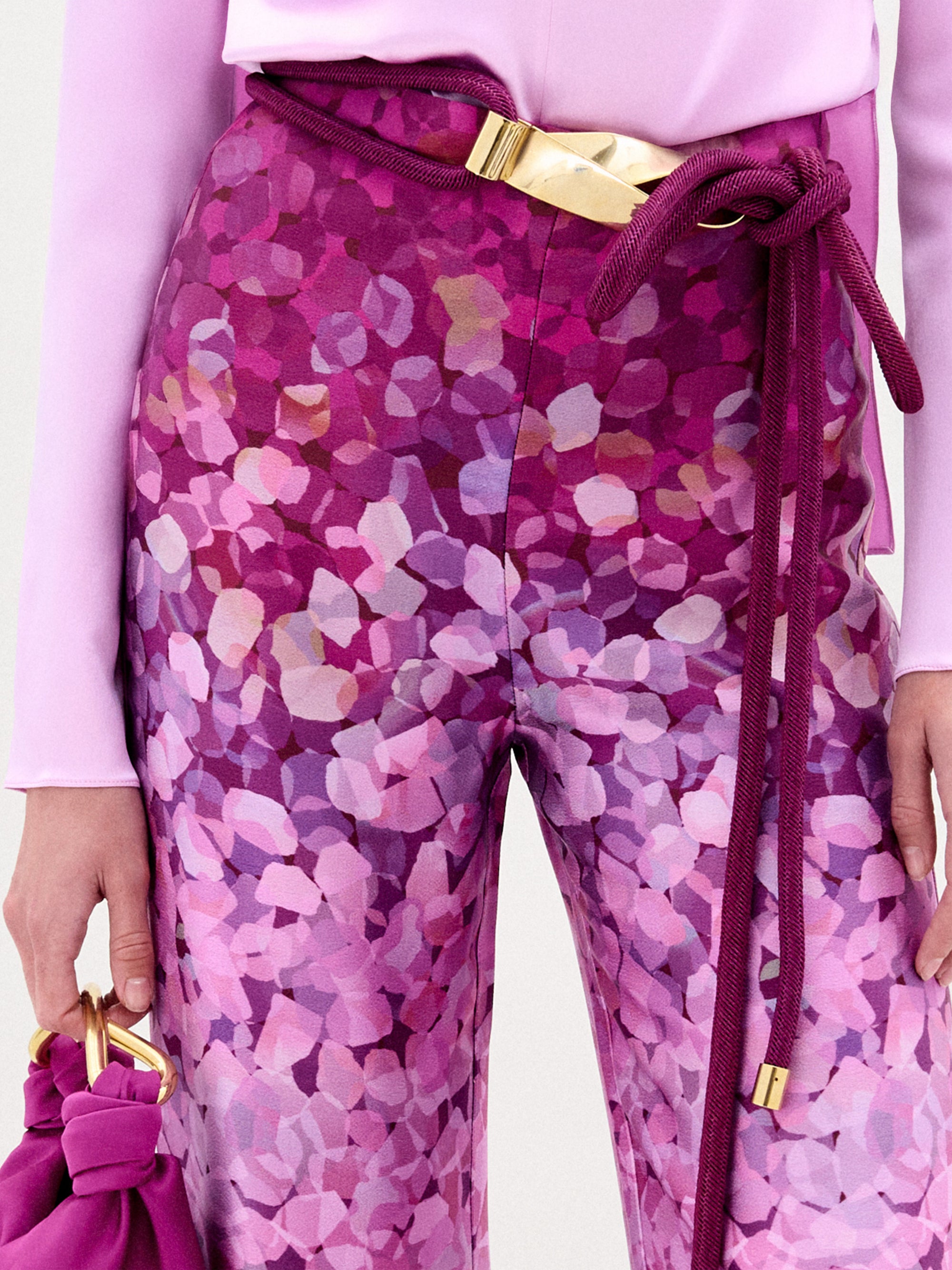 A person wearing the Como Pant—high-waisted, wide-leg pink and purple Italian fabric trousers—with a light pink long-sleeve top, magenta rope belt with gold accents, and a matching magenta bag featuring a gold handle.